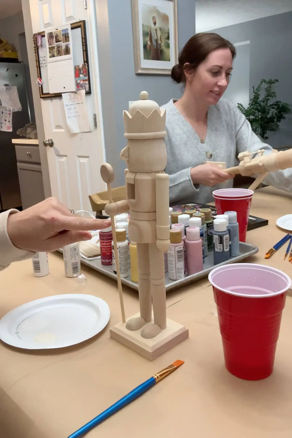 Unpainted wooden nutcracker figure standing on a table, ready to be painted. Several bottles of paint and brushes are scattered on the table, with a woman in a gray sweater working on a similar project in the background.
